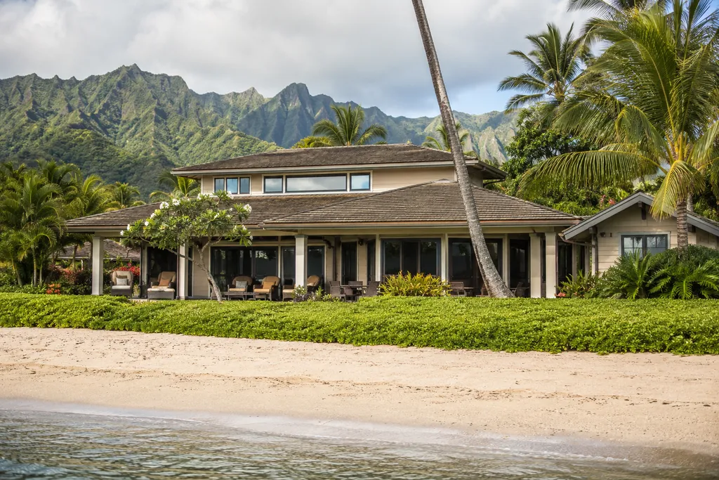 A beachfront house with a patio, surrounded by lush greenery and mountains in the distance.