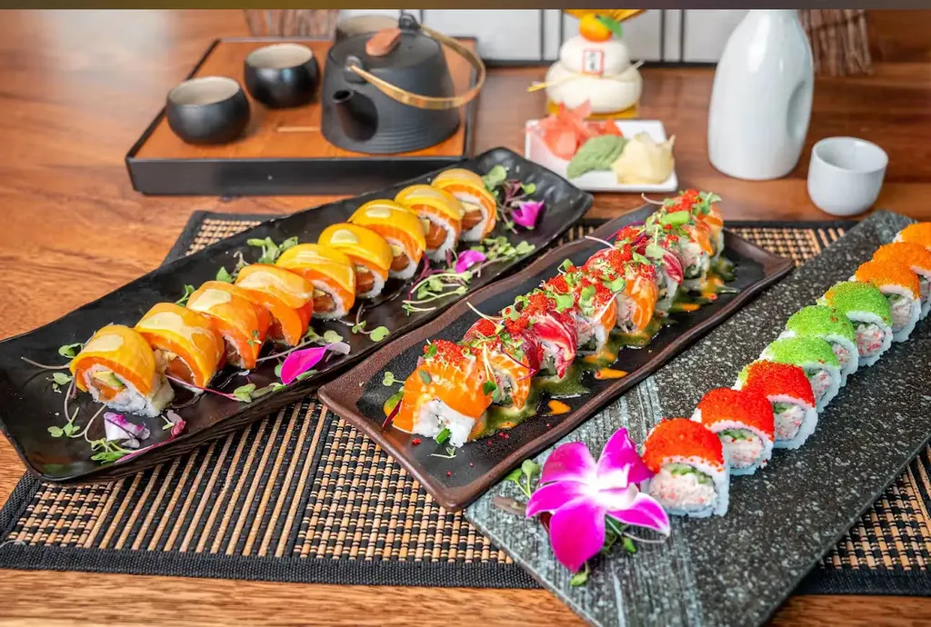 Two trays of sushi rolls on a wooden table with a flower and tea set.